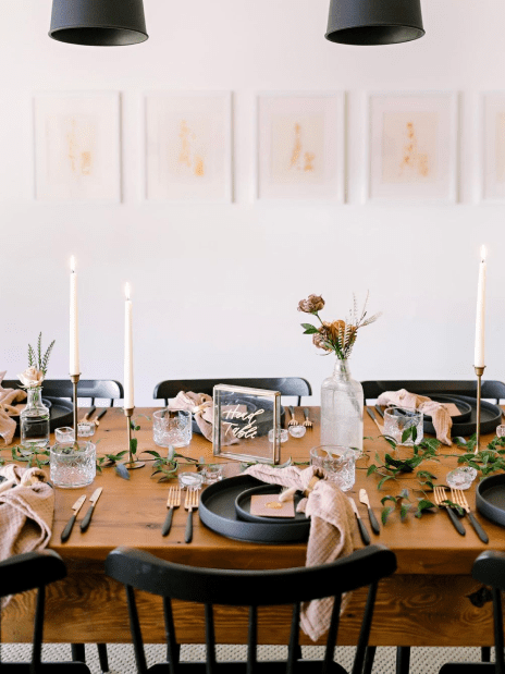 A table set with silverware and plates, candles. And flowers on the table. In front of a white wall.