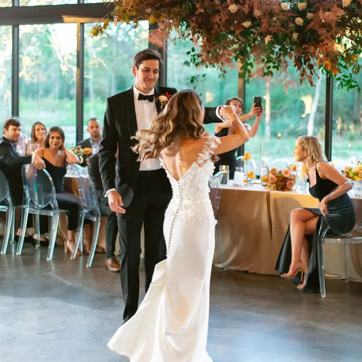 A bride and groom are dancing at their wedding. They are holding hands. There is a crowd of people in the background. One person is taking a picture with his phone camera.