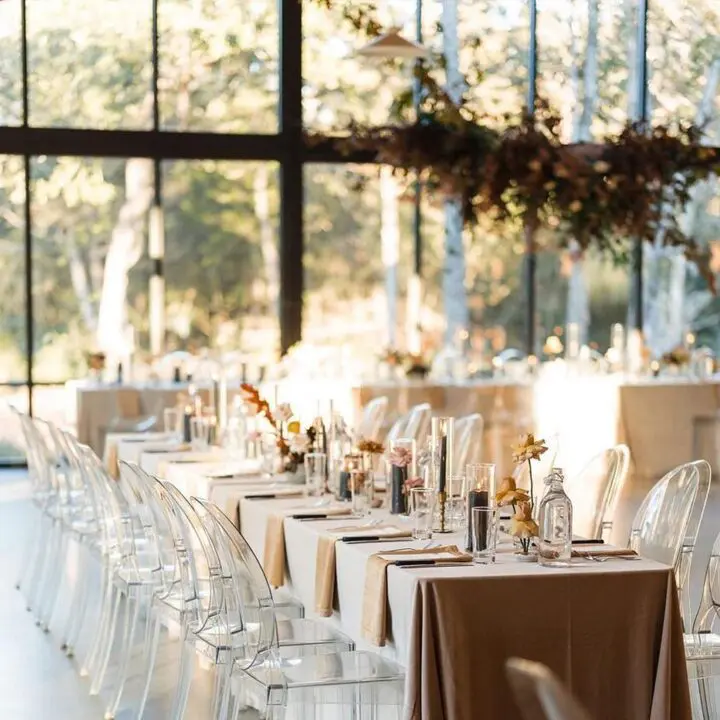 A long table with clear chairs and brown tablecloth. In front of a window. There are flowers on the tables. And trees in the background