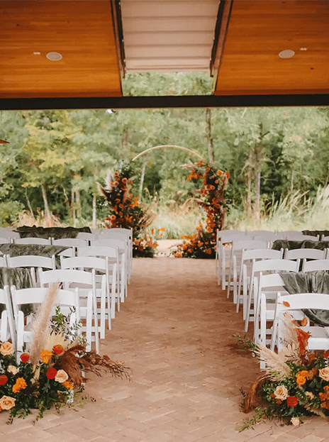 A wedding ceremony with white chairs and flowers. The room is lined with trees. There are no people in it. There is also a wooden arch that has flowers on each side.