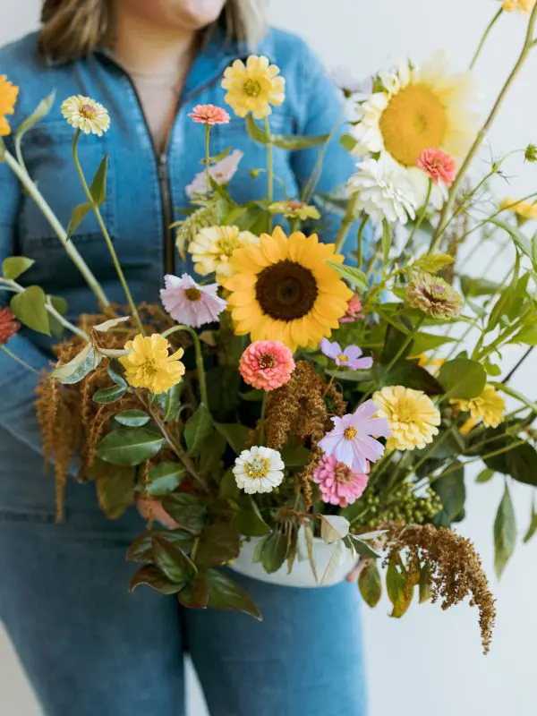 A woman holding a vase full of flowers. She is wearing blue jeans. The vase is filled with lots of different colored flowers.