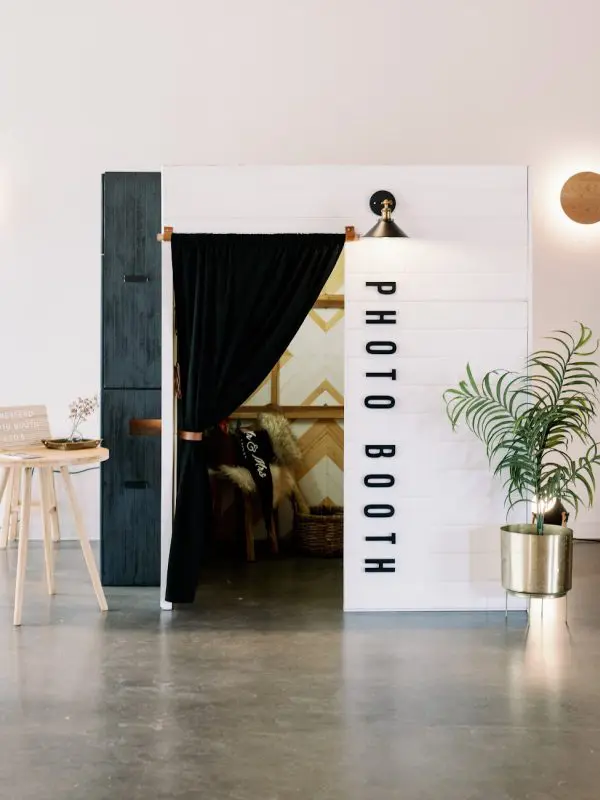 A photo booth with black curtains and white walls. There is a table, chairs, and plant in the room. It has a sign on the door that says " photo booth ".