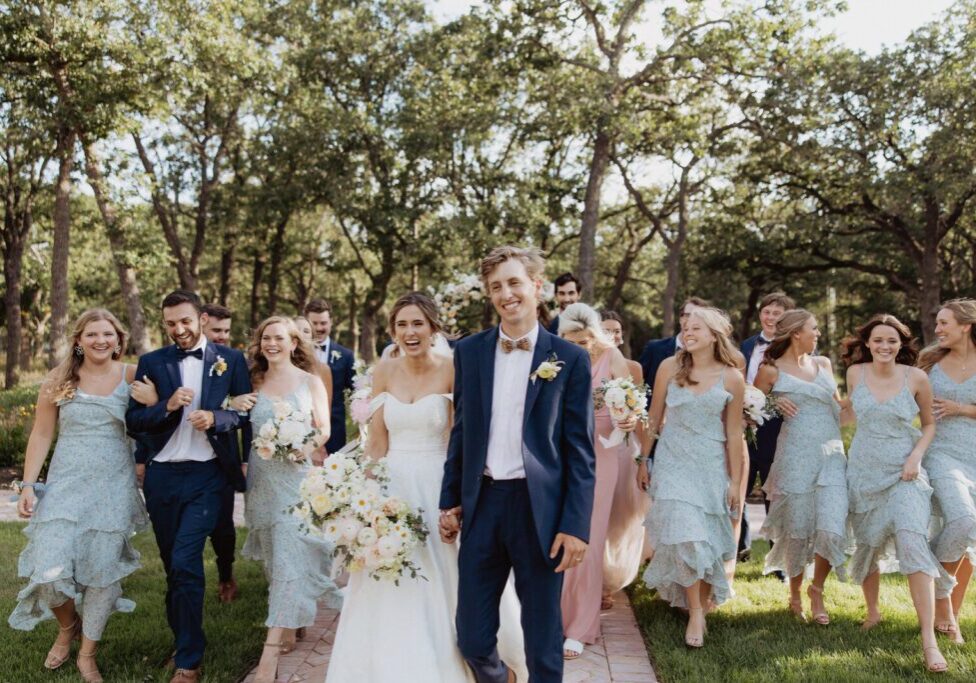 A group of people in formal wear walking down the sidewalk. They are holding flowers and smiling for the camera.